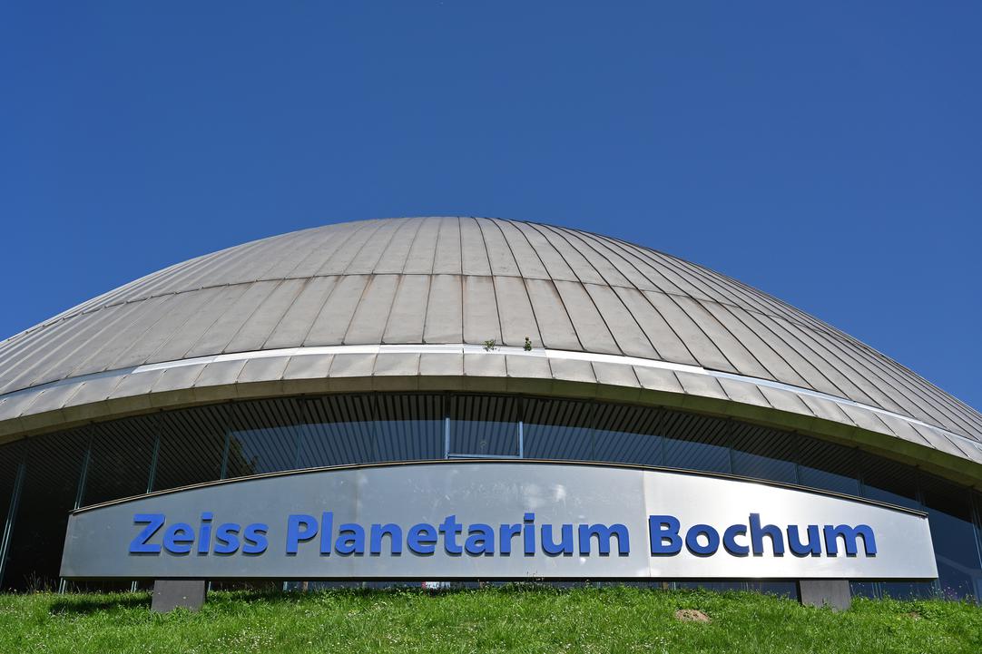 Außenaufnahme des Zeiss Planetarium Bochum. Vor dem Gebäude ist ein großes Schild auf dem „Zeiss Planetarium Bochum“ steht.