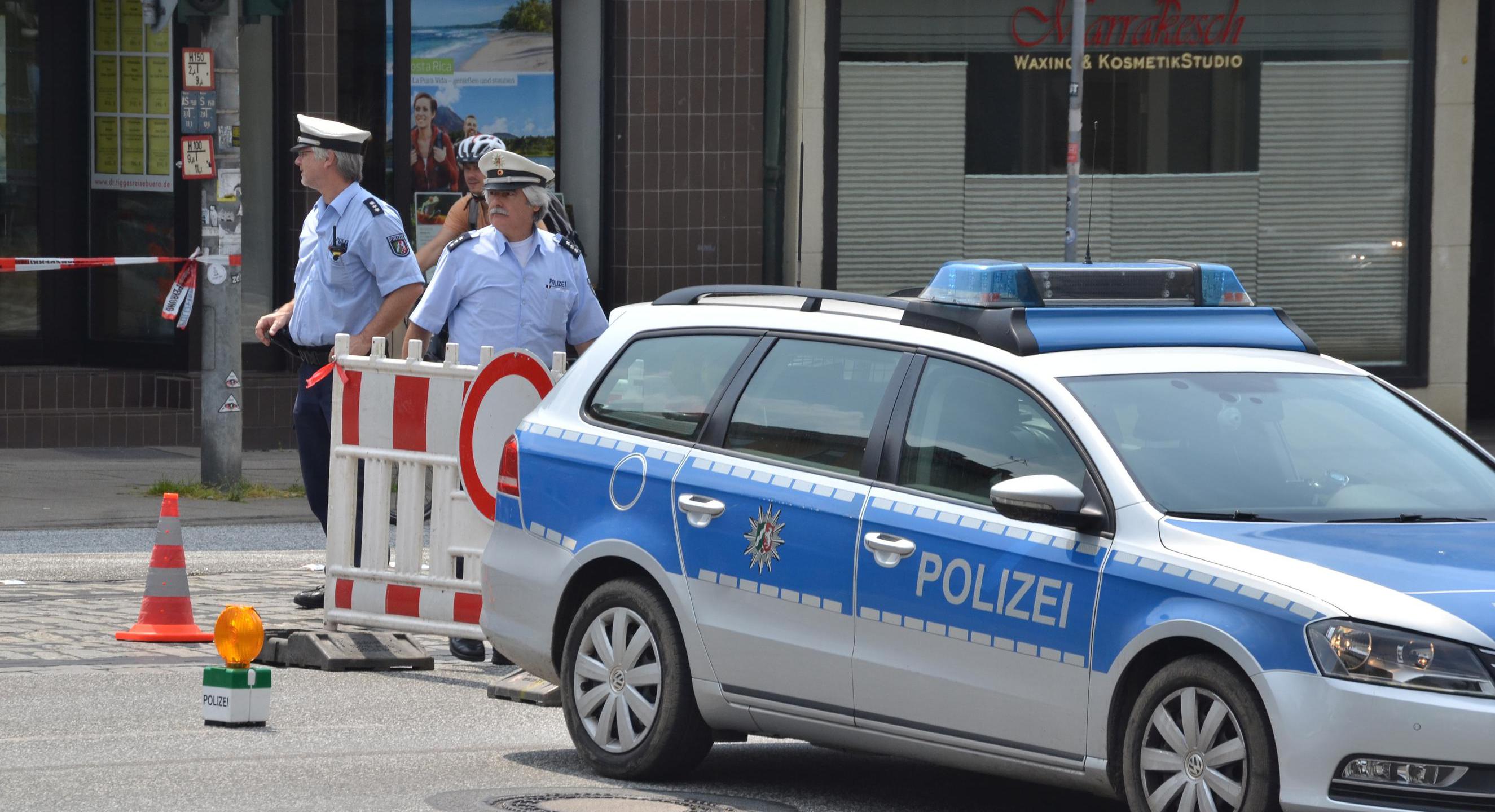 Zwei Polizisten und ein Polizeifahrzeug, mit dem eine Straße gesperrt wird.