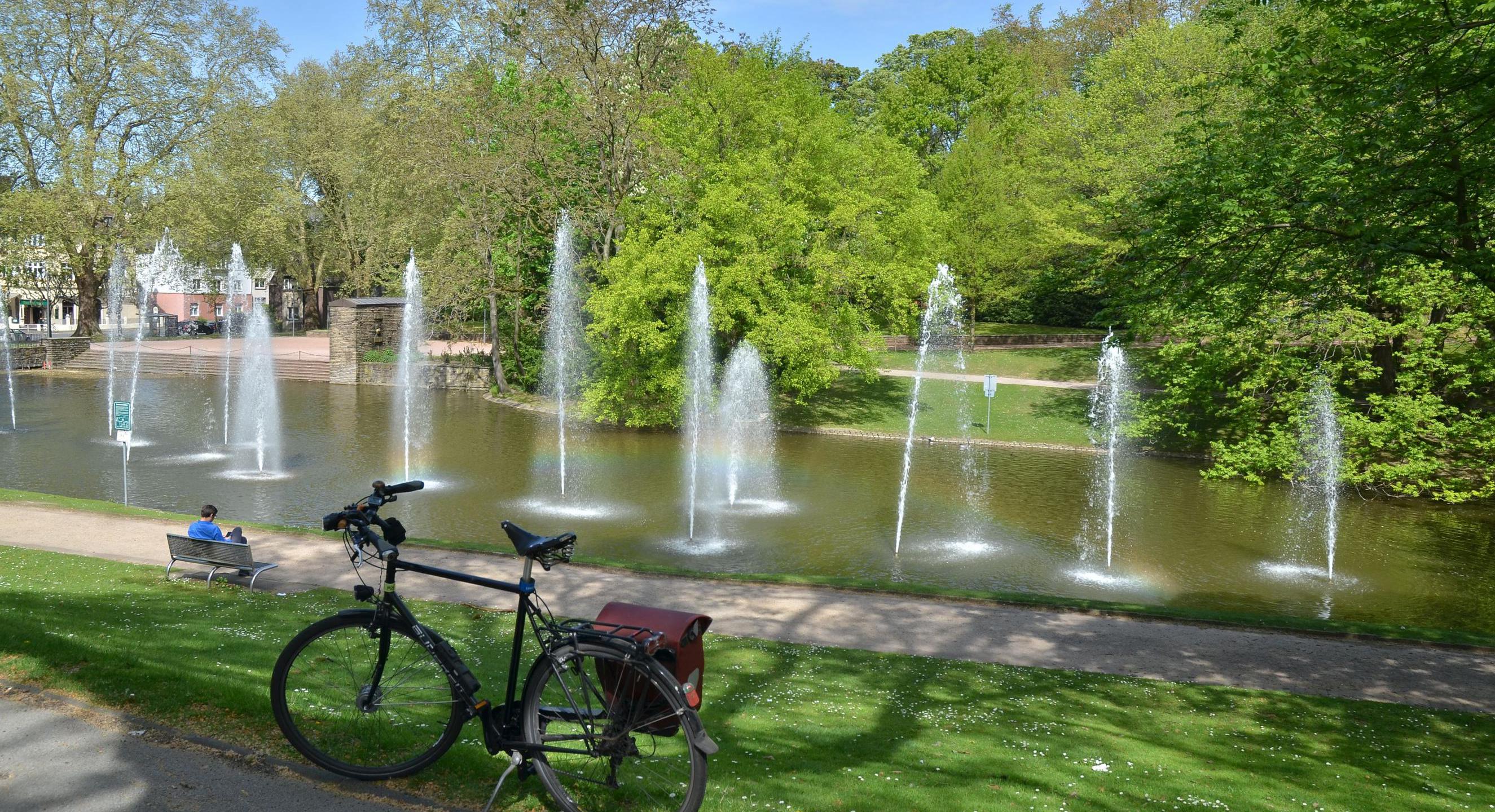 Stadtpark in Bochum - Gondelteich mit Fontänen. Im Vordergrund des Bildes steht ein Fahrrad. Ein Mann sitzt auf einer Parkbank mit Blick auf den Teich.
