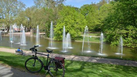 Stadtpark in Bochum - Gondelteich mit Fontänen. Im Vordergrund des Bildes steht ein Fahrrad. Ein Mann sitzt auf einer Parkbank mit Blick auf den Teich. 