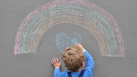 Ein Kind malt mit Straßenkreide einen Regenbogen und ein Herz auf den Asphalt.