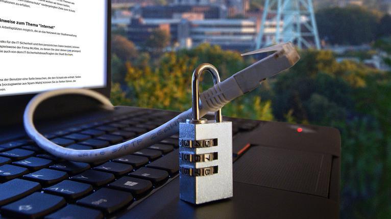 Ein Laptop mit Logo und Datenkabel in einem Vorhängeschloss - Fotomontage als Illustration zum Thema Cybersicherheit