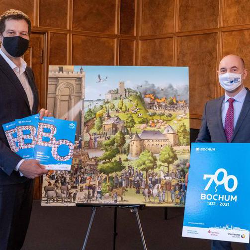 Zur Eröffnung des Festjahres hat Mario Schiefelbein (re.), Geschäftsführer von Bochum Marketing, Oberbürgermeister Thomas Eiskirch den Jubiläumsplaner übergeben. Im Hintergrund ist ein Teil des historischen Wimmelbildes zu sehen.