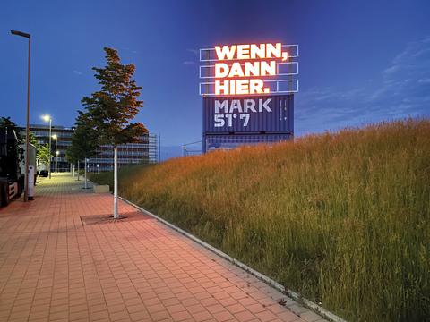 Eine leuchtende Neontafel mit der Aufschrift "Wenn, Dann, Hier.". Die Leuchttafel steht auf zwei bedruckten Containern mit der Aufschrift "MARK 51°7".