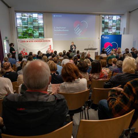 Herr Friedrich (Moderator) spricht zu den Teilnehmenden in der Aula der Evangelischen Hochschule.