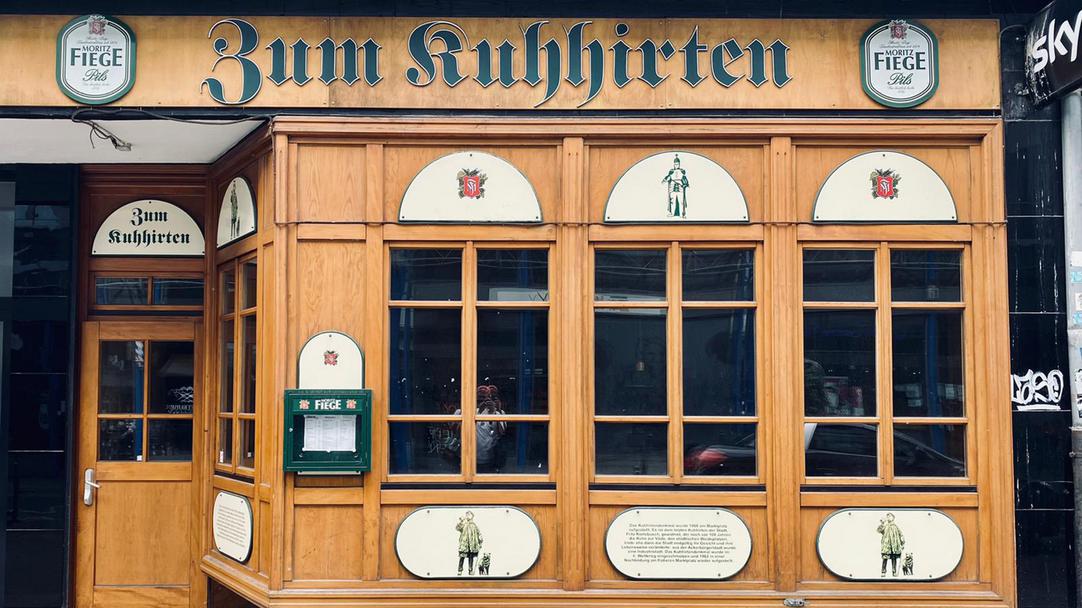 Das Bild zeigt die Traditionskneipe "Zum Kuhhirten" von außen. Links der Eingang, rechts eine hölzerne Fensterfront.
