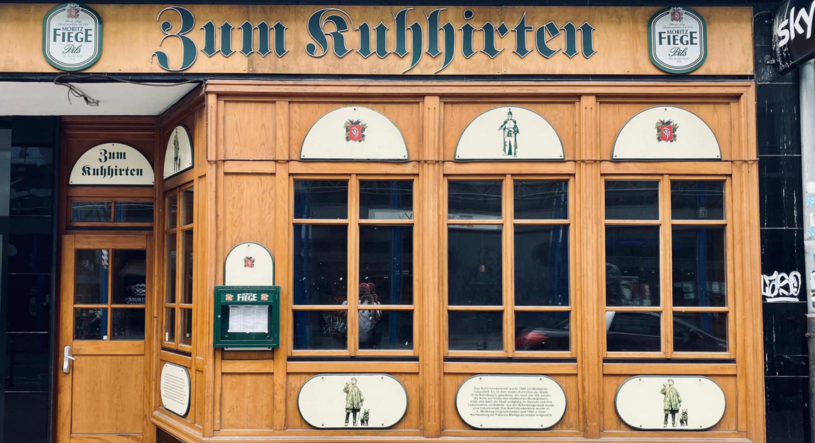Das Bild zeigt die Traditionskneipe "Zum Kuhhirten" von außen. Links der Eingang, rechts eine hölzerne Fensterfront.