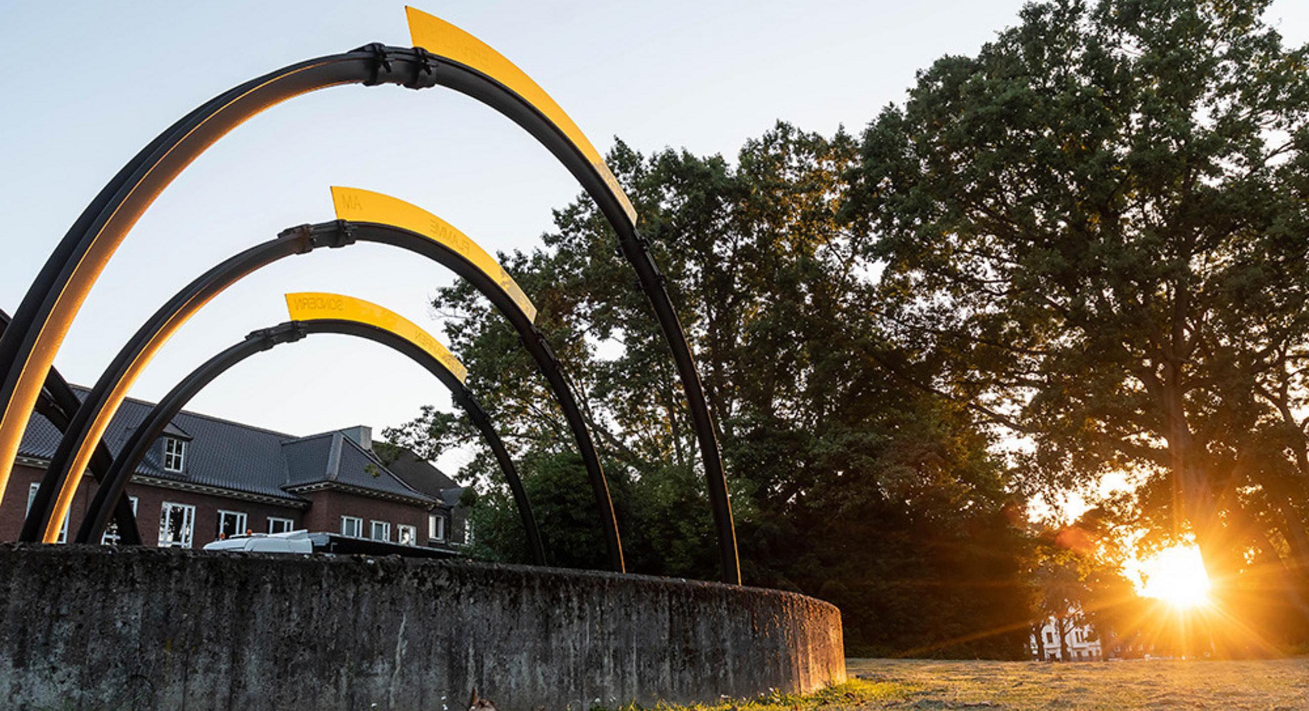 Links ist ein Kunstwerk mit einem Stein und darauf verankert drei Halbkreise zu sehen, rechts Bäume wodurch die Abendsonne scheint.