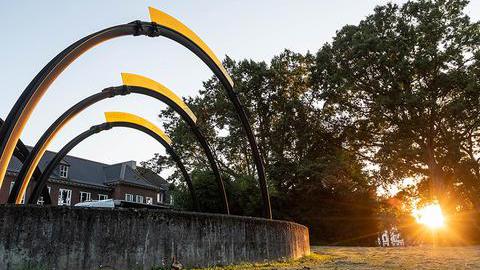 Links ist ein Kunstwerk mit einem Stein und darauf verankert drei Halbkreise zu sehen, rechts Bäume wodurch die Abendsonne scheint.