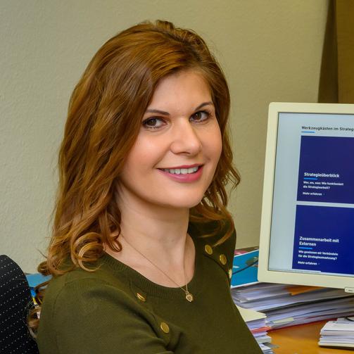 Das Bild zeigt eine ca. 40jährige Frau mit hellbraunen Haaren, einem grünen Oberteil. Sie schaut in die Kamera und sitzt am Schreibtisch.