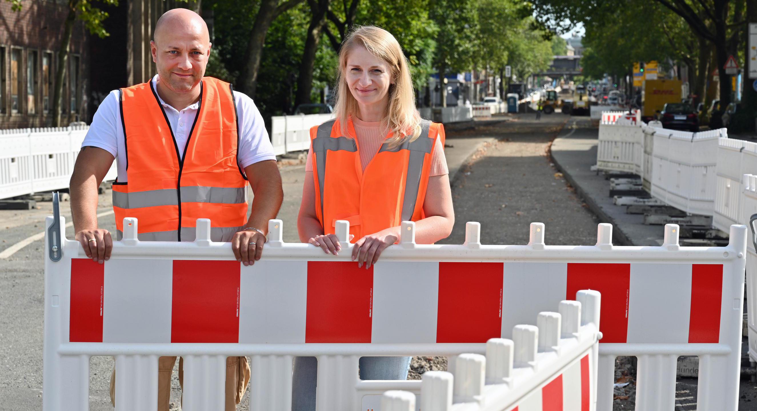 Das Bild zeigt einen Mann und eine Frau auf einer Baustelle. Sie tragen orangene Baustellenwesten.