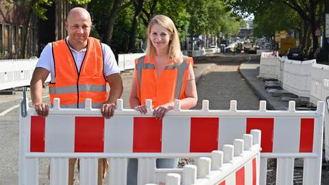 Das Bild zeigt einen Mann und eine Frau auf einer Baustelle. Sie tragen orangene Baustellenwesten.