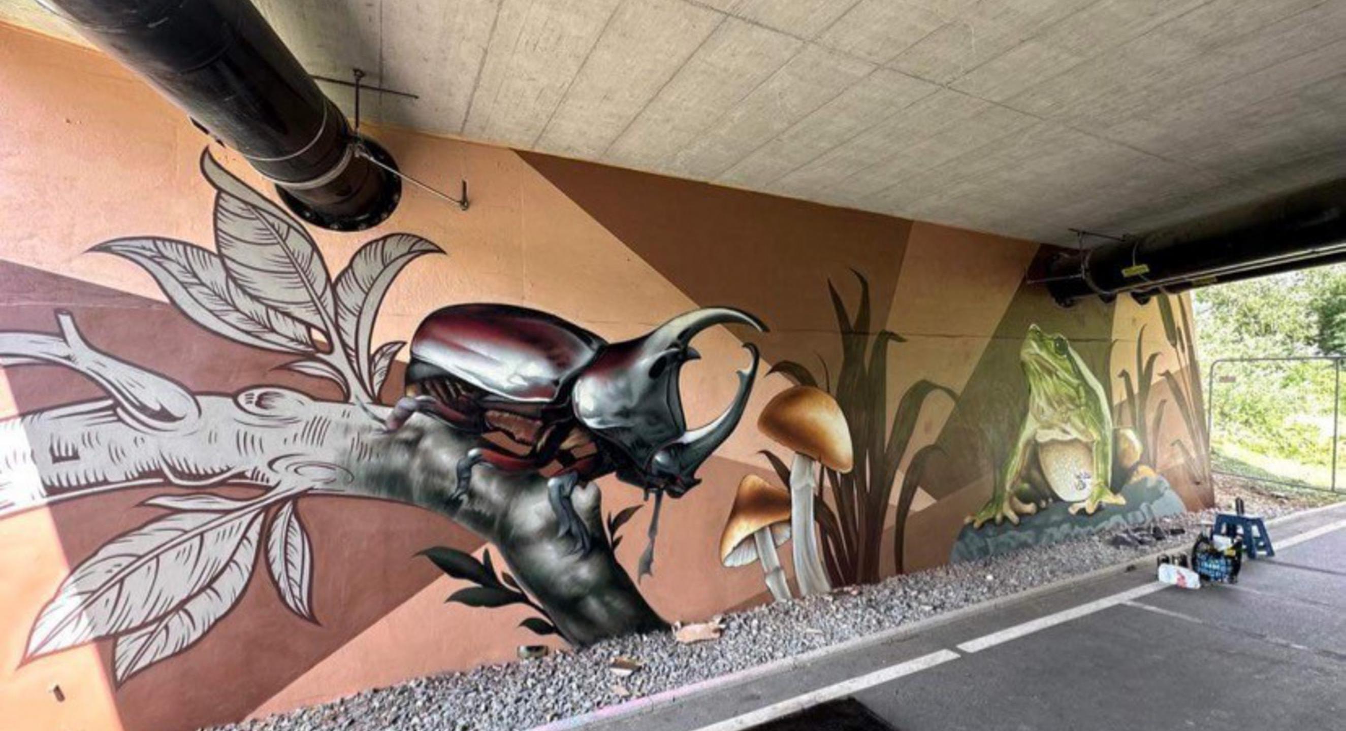 Das Bild zeigt ein Graffiti eines Käfers auf einem Ast an einer Wand unter einer Unterführung.