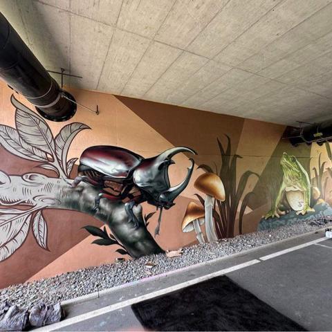 Das Bild zeigt ein Graffiti eines Käfers auf einem Ast an einer Wand unter einer Unterführung. 