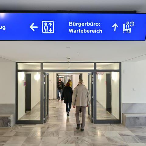 Eingangsbereich zum Bürgerbüro im Rathaus der Stadt Bochum. Zu sehen ist ein langer Flur auf dem Menschen entlanglaufen. An der Decke hängt ein digitales Display mit blauem Hintergrund.