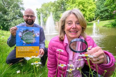 Umweltamtsleiter Philipp Heidt zeigt das Plakat zum Langen Tag der StadtNatur, während Umweltpädagogin Rita Brandenburg symbolisch Margeriten unter die Lupe nimmt.