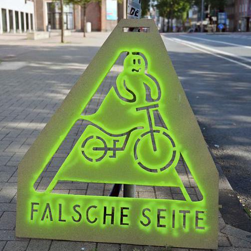 Grünes dreieckiges Warnschild mit der Aufschrift "Falsche Seite" und einem Geist auf dem Fahrrad