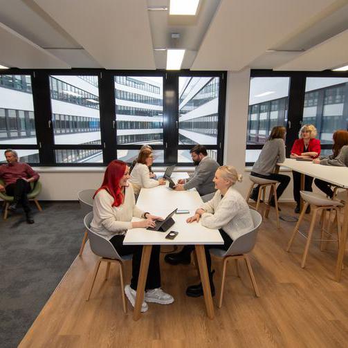 Das Bild zeigt einen modernen Arbeitsraum mit langen Tischen, an denen junge Arbeitskollegen miteinander arbeiten.