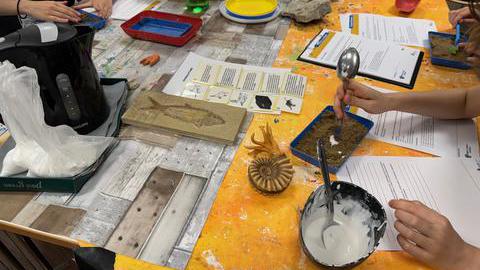 Ein gelber Arbeitstisch von oben, darauf Sand und Formen, um Fossilien selbst zu machen.