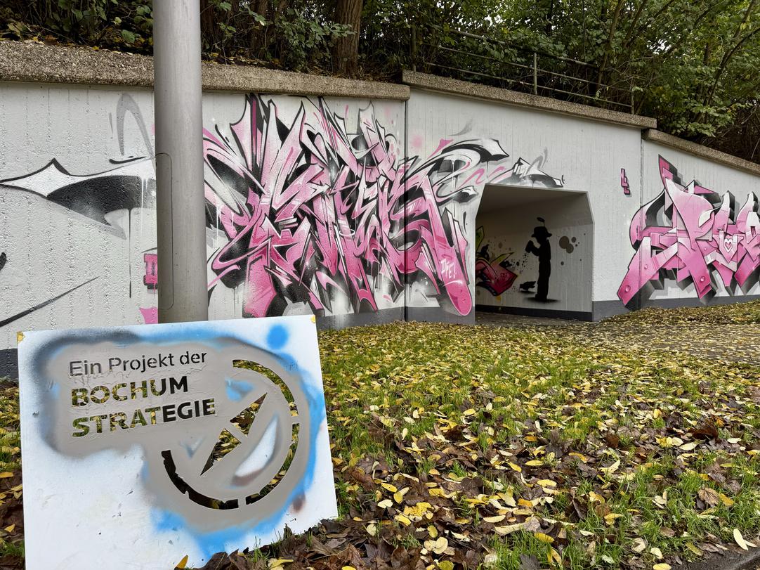 Unterführung mit pink-schwarzem Graffiti auf den Wänden und einem Schild im Vordergrund mit der Aufschrift "Ein Projekt der Bochum Strategie".