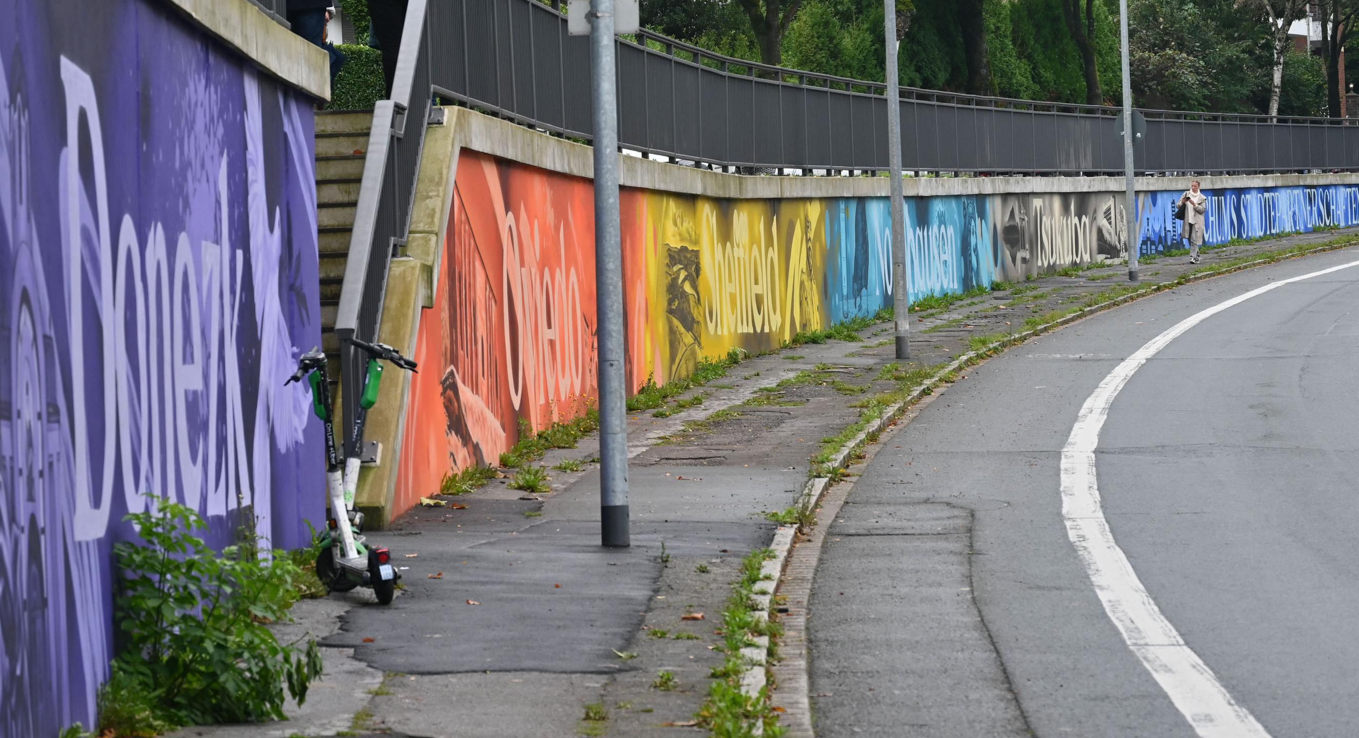 Eine Straße mit einer Kurve. Am Rand ist eine Mauer mit bunten Bildern.