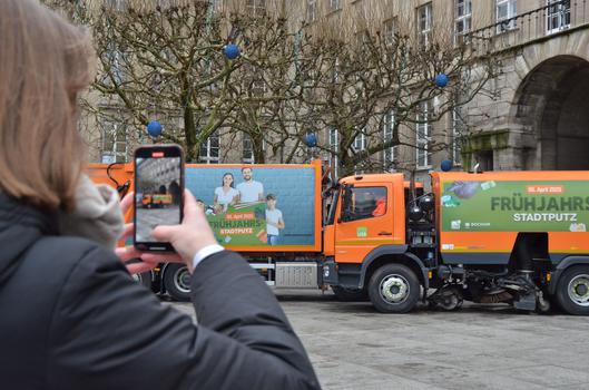 Eine Frau fotografiert mit einem Smartphone Müllwagen des USB mit dem Kampagnenaufdruck "Frühjahrs-Stadtputz".