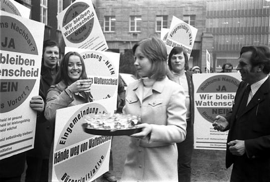 Menschen halten Protestplakate hoch und eine Frau verteilt Gläser mit Getränken an die Protestierenden.