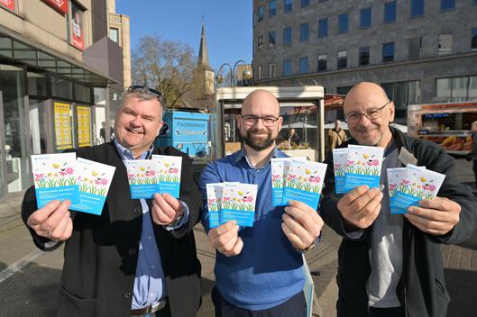 Jörg Czwikla vom Umweltausschuss, Philipp Heidt vom Umweltamt und Stefan Welzel von der Biologischen Station verteilen Tütchen mit einer Wildblumenmischung im Rahmen von „Bochum blüht und summt“ auf dem Wochenmarkt in Bochum