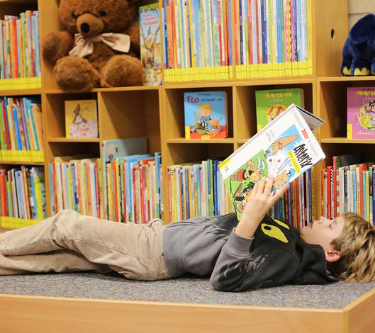 Ein Kind liegt auf dem Rücken vor einem Bücherregal und liest ein Buch.