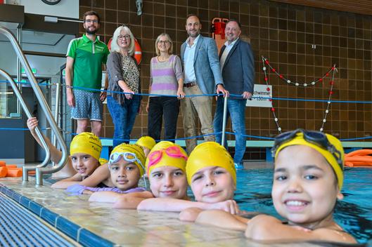 Auf dem Gruppenfoto sind im Vordergrund Kinder im Schwimmbecken und im Hintergrund stehen Erwachsene am Beckenrand.