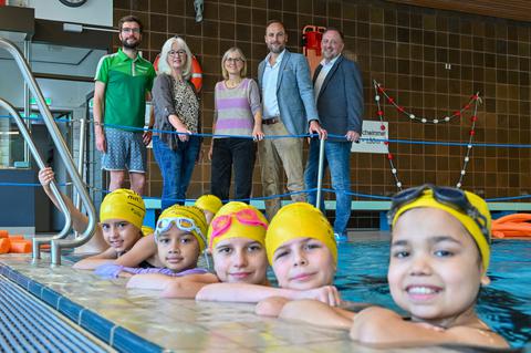 Auf dem Gruppenfoto sind im Vordergrund Kinder im Schwimmbecken und im Hintergrund stehen Erwachsene am Beckenrand.