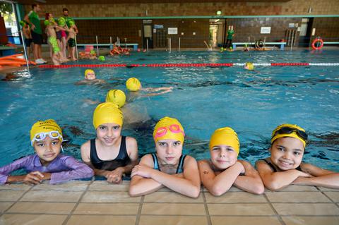 Kinder lehnen am Beckenrand und schauen in die Kamera. Im Hintergrund findet Schwimmunterricht statt.