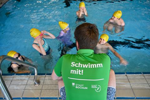 Es ist ein Mann von hinten zu sehen, der mit den Kindern im Wasser spricht.