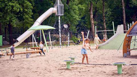 Ein Bild eines Spielplatzes mit Rutsche, Wippen und einem Seil zum Balancieren, auf dem ein Mädchen steht.