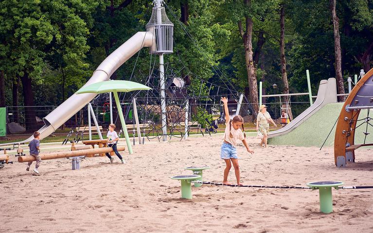 Ein Bild eines Spielplatzes mit Rutsche, Wippen und einem Seil zum Balancieren, auf dem ein Mädchen steht.