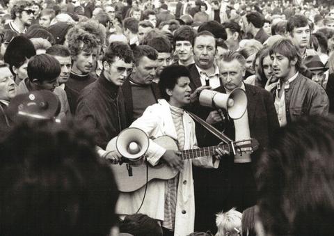 Fasia Jansen steht mit einer Gitarre vor einem Megaphon. Hinter ihr befindet sich eine Menschenmenge.
