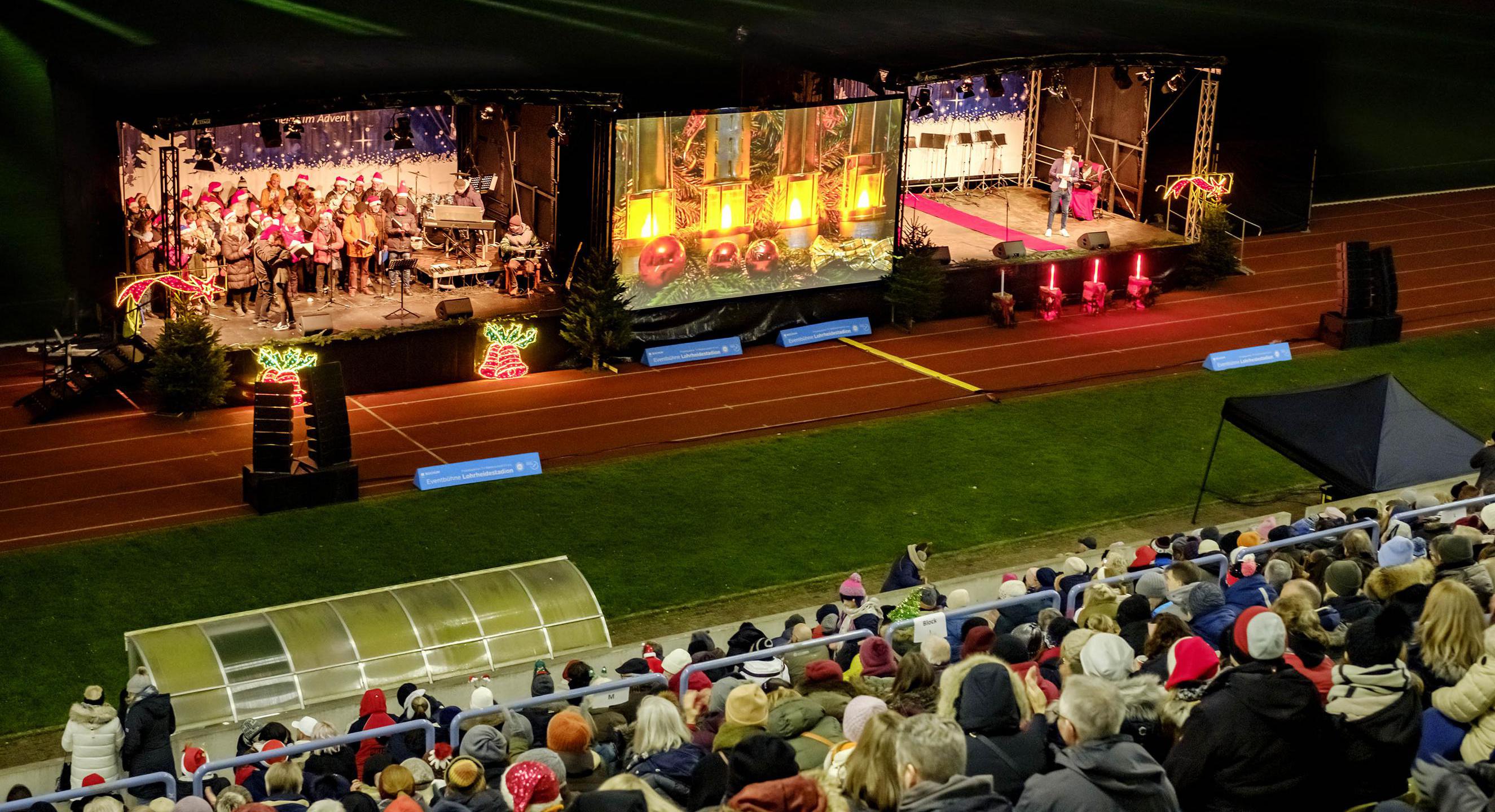 Zwei Bühnen in einem Stadion. Auf den Bühnen sind zwei Chorgruppen zu sehen. Davor sitzen Menschen auf Tribünen.