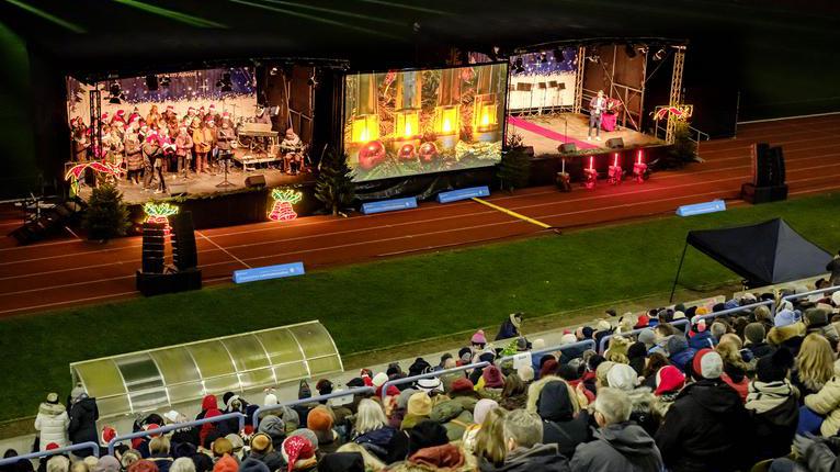 Zwei Bühnen in einem Stadion. Auf den Bühnen sind zwei Chorgruppen zu sehen. Davor sitzen Menschen auf Tribünen.
