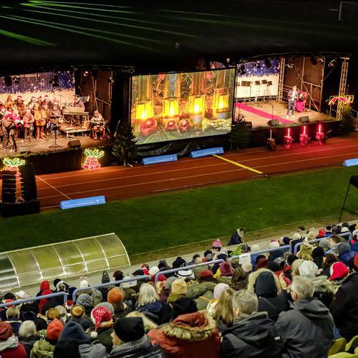 Zwei Bühnen in einem Stadion. Auf den Bühnen sind zwei Chorgruppen zu sehen. Davor sitzen Menschen auf Tribünen.