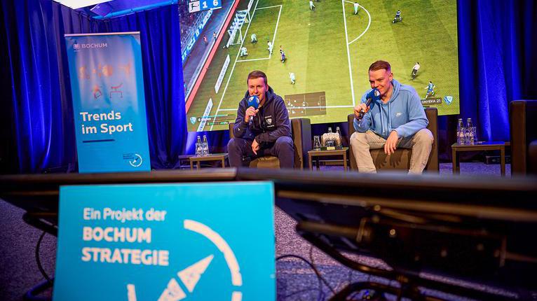 Auf dem Bild sitzen zwei junge Männer mit Mikrofonen in der Hand auf einer Bühne und kommentieren ein Fußballspiel.