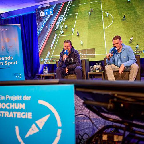 Auf dem Bild sitzen zwei junge Männer mit Mikrofonen in der Hand auf einer Bühne und kommentieren ein Fußballspiel.