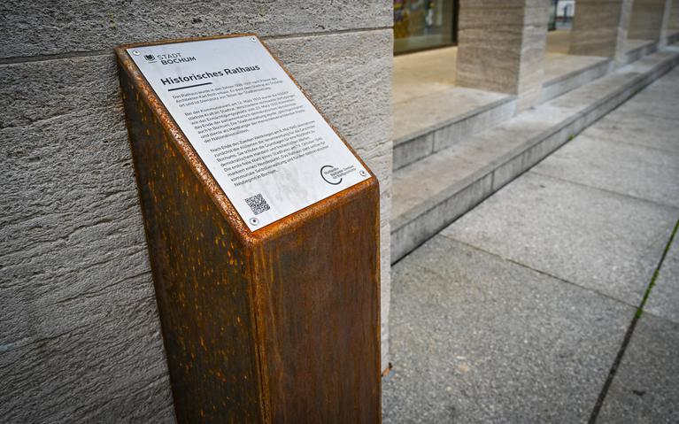 Eine rostbraune viereckige Stele vor dem Bochumer Rathaus. Auf ihr ist eine metallene Platte angebracht mit Informationen zum Historischen Rathaus. Die Logos der Stadt Bochum und des Stadtarchivs sowie ein QR-Code sind ebenfalls auf der Metallplatte zu sehen.