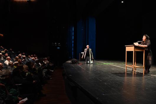 Ein Mann kniet auf der Bühne und eine Frau liest an einem Podium etwas vor.