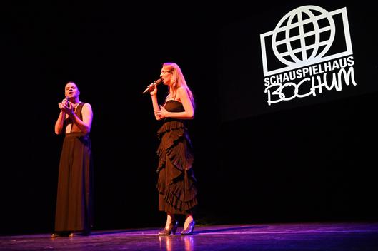 Zwei Personen stehen mit Mikrofonen in der Hand auf einer Bühne.