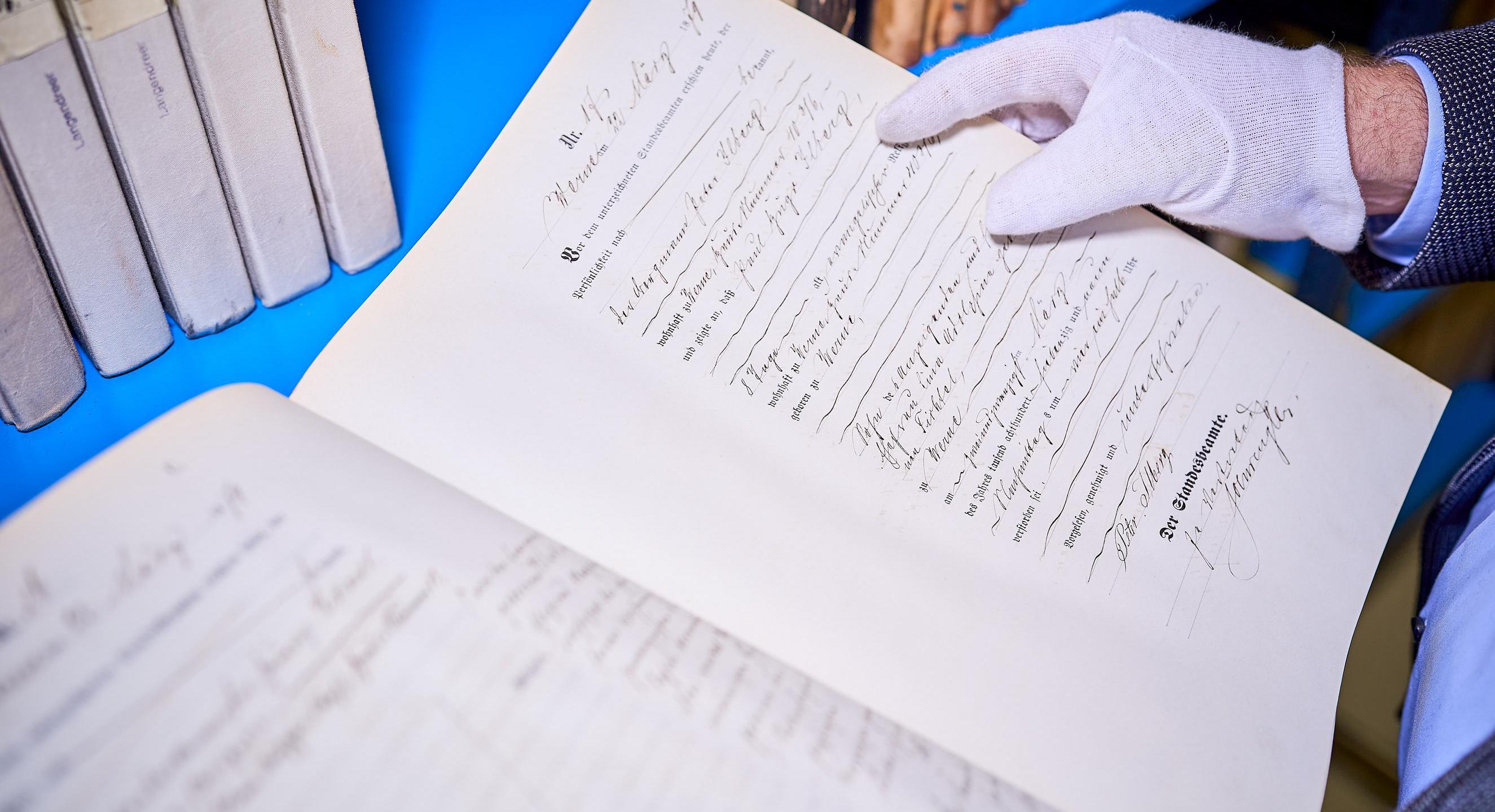 Ein altes leicht gelbliches Dokument in verschnörkelter Schrift. Die Schrift ist teils gedruckt und teils mit der Hand geschrieben. Unten steht in größerer Schrift "Der Standesbeamte". Weiße Baumwollhandschuhe halten das Dokument fest. Im Hintergrund sind alte Bücher zu sehen.