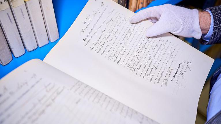 Ein altes leicht gelbliches Dokument in verschnörkelter Schrift. Die Schrift ist teils gedruckt und teils mit der Hand geschrieben. Unten steht in größerer Schrift Der Standesbeamte. Weiße Baumwollhandschuhe halten das Dokument fest. Im Hintergrund sind alte Bücher zu sehen.