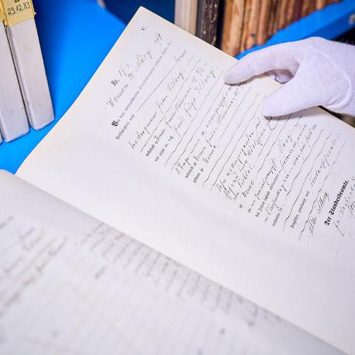 Ein altes leicht gelbliches Dokument in verschnörkelter Schrift. Die Schrift ist teils gedruckt und teils mit der Hand geschrieben. Unten steht in größerer Schrift Der Standesbeamte. Weiße Baumwollhandschuhe halten das Dokument fest. Im Hintergrund sind alte Bücher zu sehen.