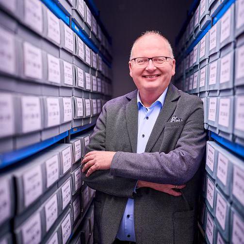 Kai Tawe steht zwischen zwei Regalen gefüllt mit Boxen. Er verschränkt die Arme und lächelt in die Kamera. Er trägt einen Anzug und eine Brille.
