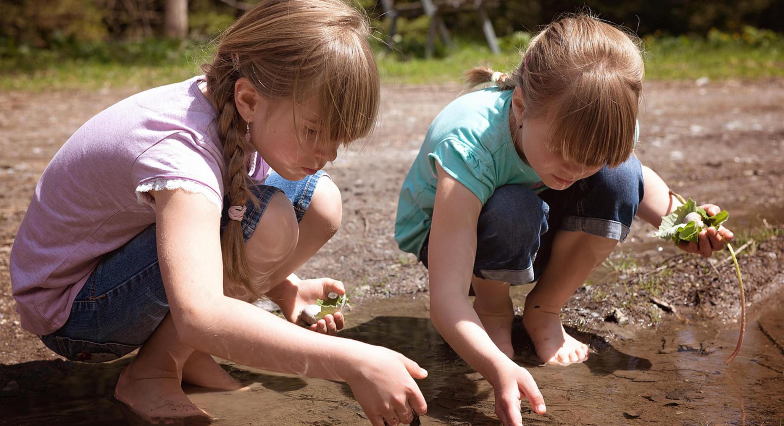 Zwei Kinder Hocken Barfuß in einer Pfütze im Wald. Sie tragen kurze Hosen und T-shirts. In der linken Hand halten beide Blätter. Mit der rechten Hand wühlen sie durch die Pfütze.