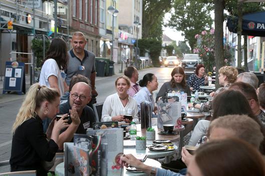 Personen sitzen an Tischen in einem Straßencafé und unterhalten sich.